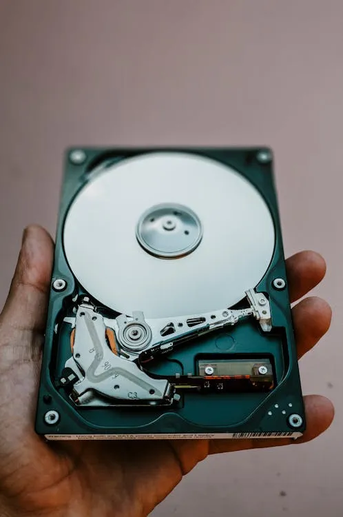 An opened hard drive disk held in a hand, with the read/write head visible against the platter.